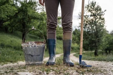 Metal kovası ve tırmıkla toprak yolda dikilen kauçuk çizmeli bilinmeyen kadının bacaklarına yaklaş - tarım ve sürdürülebilir yaşam konsepti
