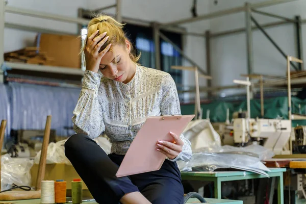 Portrait of adult caucasian woman entrepreneur sitting at factory holding clip chart documents reading reports holding her head - Sad female depressed about bad business results - recession and crisis