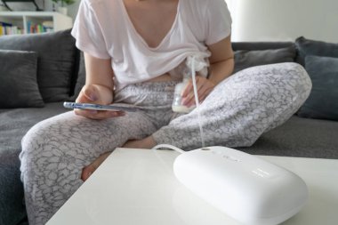 Bebeği için süt pompalayan ve cep telefonu uygulamasında, ebeveynlik ve annelik kavramını takip eden kimliği belirsiz beyaz bir kadının karnını kapatın.