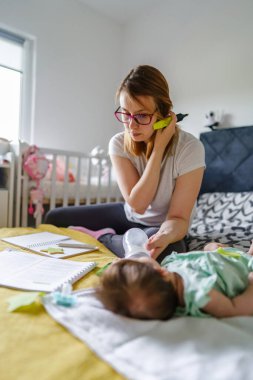 Bin yıllık beyaz kadın annenin evde ders çalışırken yatakta oturması ve yeni doğmuş bebeğini emzirme formülü ile beslemesi - eğitim kavramı gerçek insanlar