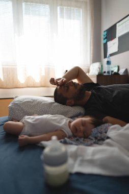 Yorgun bir baba, üç aylık bebeğinin yanında yatarken evde uyuyor. Bebek bakıcılığı konseptini seçici bir şekilde beslerken el ele tutuşuyor.
