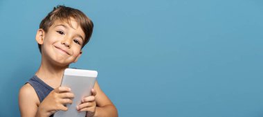 Dört yaşında, mavi arka plan stüdyosunun önünde dikilip dijital tablet kullanarak kameraya gülümseyen küçük beyaz bir çocuğun ön görüntüsü.