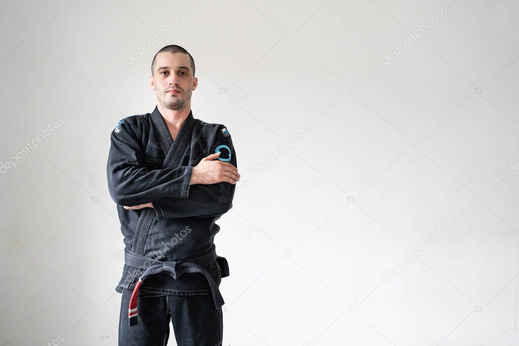 Front view of adult male athlete bjj brazilian jiu jistu black belt standing in front of white wall in kimono gi with copy space looking to the camera