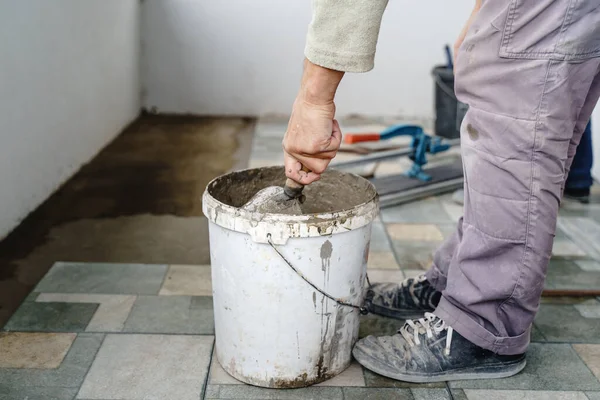 Side view on bucket with adhesive construction glue cement and unknown craftsmen holding tool in hand preparing material for ceramic tiles