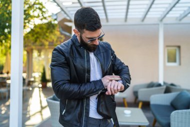 Yetişkin beyaz bir adam güneşli bir sonbahar gününde dışarıda kafede beklerken kol saatini kontrol ediyor. Randevu ya da buluşma saatini ya da geç saatte buluşmayı bekliyor. Sabır konsepti kopyalama alanı.