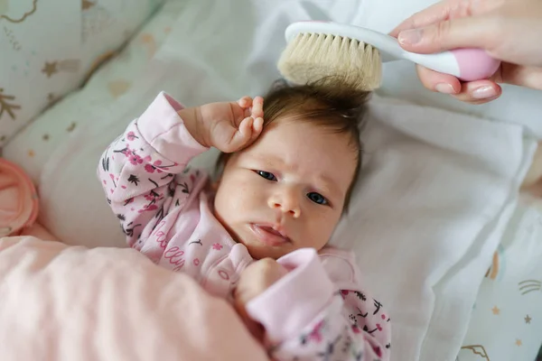 Top view on small caucasian baby two months old lying in bed while unknown woman mother hand is taking care of her hair with brush brushing