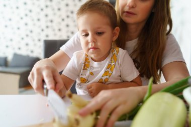 Anne ve oğlunu ön planda otururken yemek hazırlarken küçük oğluyla birlikte beyaz bir kadın sebze doğramak için zaman harcıyor.