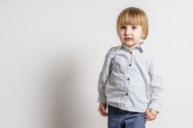 Beyaz duvarın önünde duran küçük sarışın beyaz çocuğun yarım boy portresi. Yanında fotokopi alanı olan küçük çocuk stüdyosu resmi.