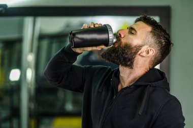 Siyah kapüşonlu genç beyaz erkek sporcunun ön manzara portresi. Spor salonunda elinde protein takviyesi olan siyah saç ve sakal fotokopi içeceği.