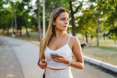 Yazın ya da sonbahar günü parkta beklerken cep telefonu kullanan genç beyaz kadın beklerken kaşlarını çatıyor.