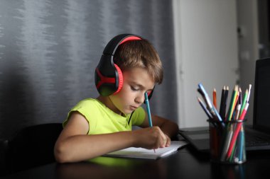 Bilgisayarda kulaklıkla ders dinleyen ve not defterine yazı yazan tatlı bir okul çocuğu. Eğitim ve okula dönüş.