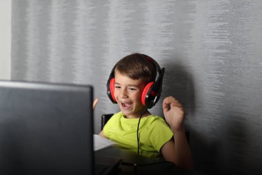 Dişleri olan, gülümseyen ve elleri havada, evdeki bilgisayar ekranına bakan çok mutlu bir çocuk. Çocuk kafasına kulaklık takıyor ve müzik dinliyor.
