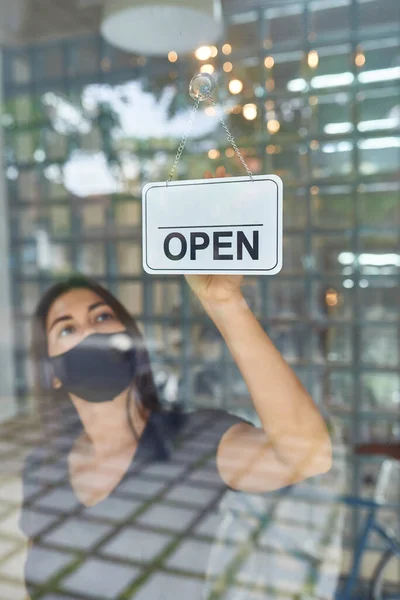 Beautiful European girl in a mask hangs a sign on a glass door of a clothing store (open) after a cowid-19 epidemic