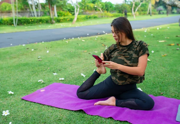 A beautiful Caucasian girl practices a yoga lesson online, through a phone in an outdoor garden during quarantine.