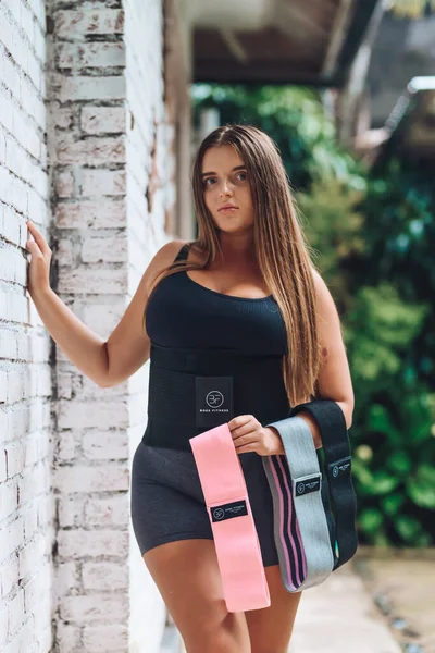 Attractive young overweight woman in active clothes, choosing a healthy lifestyle. Body shape and activity with fitness belt for sweat and resistance band.