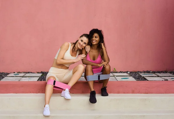 Fitness girls in stylish sports suits do exercises with rubber bands on the background of a light pink wall. Sports women are engaged in sitting on the street.
