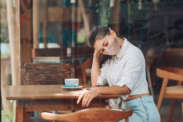 A sweet girl of Caucasian appearance in a silk face mask, sitting at a table. Photograph through transparent glass