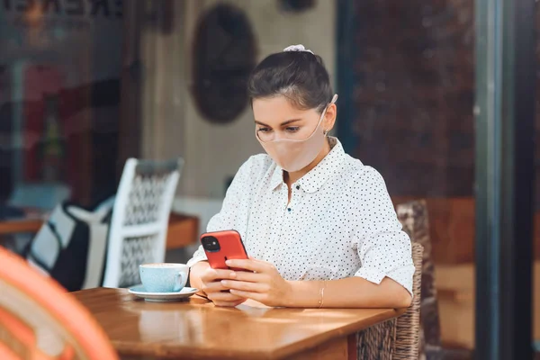 A sweet girl of Caucasian appearance in a silk face mask, sits at a table and communicates on the phone. Photograph through transparent glass