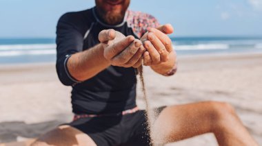 Genç adam bir sörf tahtasıyla deniz kıyısında oturur ve kumla oynar. Kuma ve ellere odaklan.