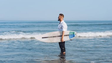 Beyaz tişörtlü genç bir Avrupalı elinde sörf tahtasıyla gezintiye çıkmak üzere..