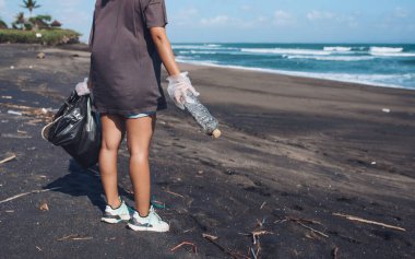Genç bir kız denizin arka planında boş bir plastik şişe ve bir çöp torbası tutuyor. Vurulmuş mantar.