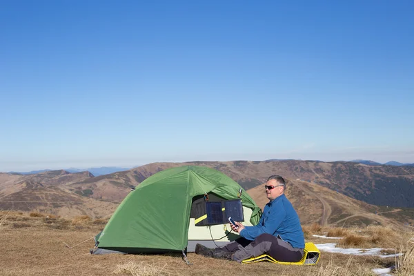 O painel solar ligado à tenda. O homem sentado ao lado das cargas do telefone móvel do sol . — Fotografia de Stock