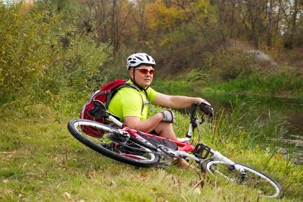 Young bicyclist in helmet sitting on a green grass near the bicycle — Stock Photo, Image