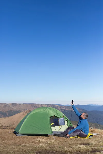 テントの中に接続されている太陽電池パネル。太陽から携帯電話料金の横に座っている男. — ストック写真
