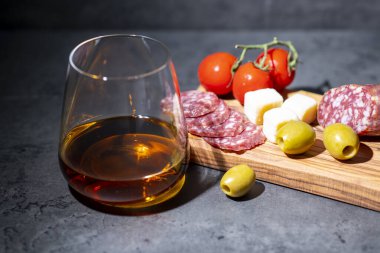 Bir bardak viski ve atıştırmalık sosis, zeytin ve domatesler koyu gri çimento arka planda.