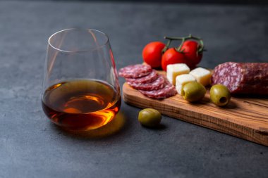 Bir bardak viski ve atıştırmalık sosis, zeytin ve domatesler koyu gri çimento arka planda.