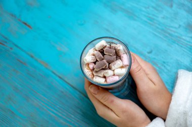 Şeffaf bardakta kapuçino, marşmelov ve mavi ahşap arka planda dişi ellerinde kakao tozu.