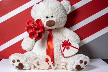 Kırmızı kravatlı, çiçekli ve kalp şeklinde hediye kutusu olan oyuncak ayı.