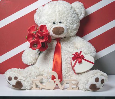 Kırmızı kravatlı, çiçekli ve kalp şeklinde hediye kutusu olan oyuncak ayı.