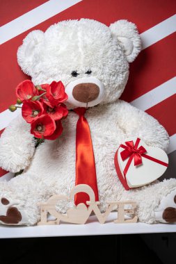 Kırmızı kravatlı, çiçekli ve kalp şeklinde hediye kutusu olan oyuncak ayı.