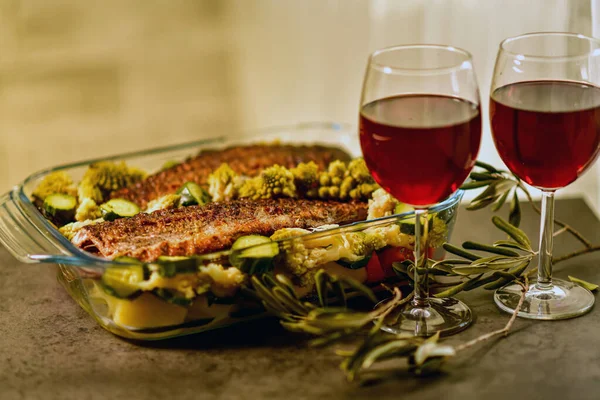 two glasses of red wine and a large glass dish with baked fish and vegetables