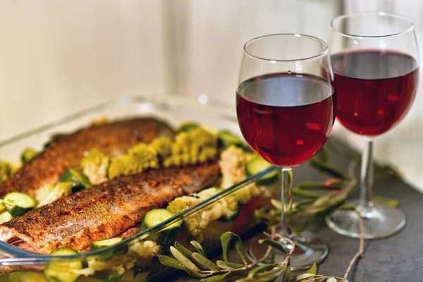 two glasses of red wine and a large glass dish with baked fish and vegetables