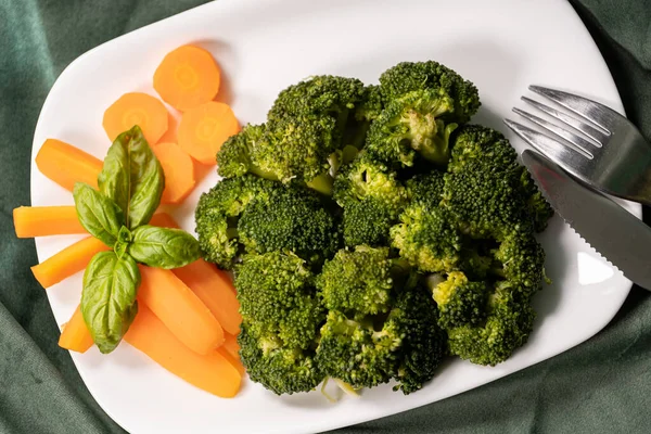 healthy food, boiled broccoli, carrots and basal leaf on a white plate on a dark green cloth