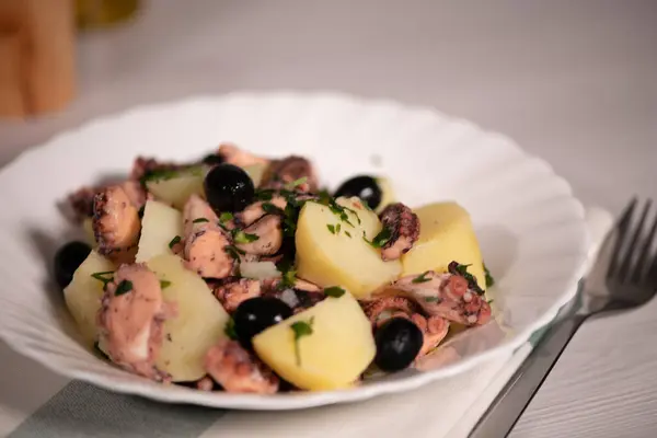 Açık gri bir masada beyaz tabakta patates ve zeytinli ahtapot salatası.