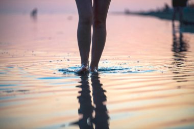 Gün batımında sahilde bir kız suyun üzerinde yürür sadece bacakları görünür.