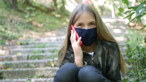 Nemocné dítě nosí ochrannou masku kvůli koronaviru pandemie, Kid mluví na Smartphone v parku, Dívka komunikuje na chytrém telefonu — Stock video