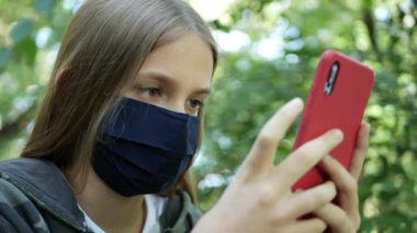 Koronavirüs salgını nedeniyle koruyucu maskeli hasta kız, Parktaki Smartphone 'da internete bakan çocuk, Akıllı Telefon Oynayan çocuk