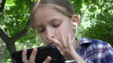 Çocuk parkta akıllı telefon oynuyor, kulaklıklı kız Bench Browsing Internet 'te oturuyor, açık havada müzik dinliyor.
