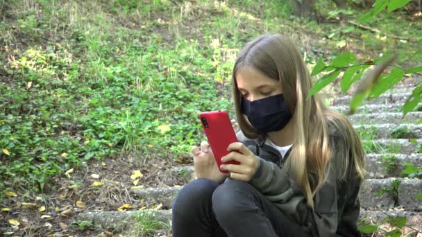 Ziek meisje met beschermende masker als gevolg van Coronavirus pandemie, Kid Browsing Internet op Smartphone in Park, Kind spelen Smart Phone — Stockvideo