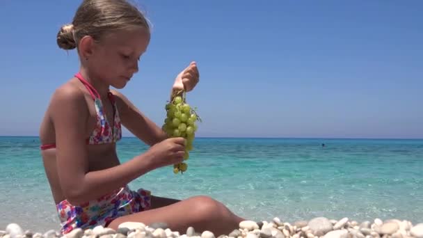 Діти їдять виноград на пляжі, кід їсть фрукти на узбережжі, блондинка маленька дівчинка грає на узбережжі, морські хвилі в літній відпустці. — стокове відео