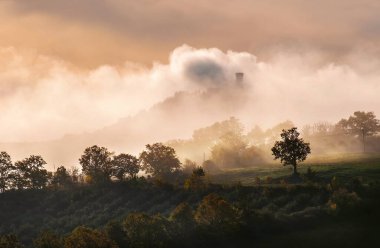 Rocca di Radicofani ana kulesi sabah sisiyle kaplı. Güneş ışığı, çimlerin ve tarlaların üzerinden akan kıvırcık bulutları kaplıyor. Tuscany tepelerinde ışık gölgeleri oluşturuyor..