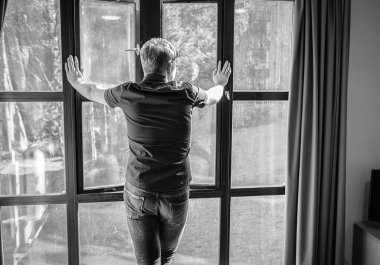 Düşünceli bir genç adam bütün pencerenin yanında duruyor ve sokaktan dışarı bakıyor ve bir şeyler düşünüyor. Kişisel izolasyon, karantina ve zihinsel sağlık konsepti..