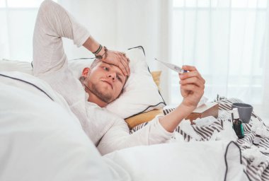 Tired and unhealthy looking at a medical thermometer as lying in a cozy home bed beside a lot of used paper tissues and medicines on the blanket. Season virus flu and a home quarantine concept image.