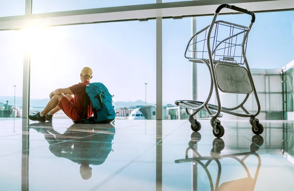 Samotny Podróżnik Siedzący Lotnisku Podłodze Terminalu Pustym Wózkiem Lotniskowym Patrzący — Zdjęcie stockowe