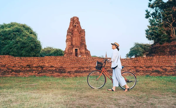 Woman walking with a bicycle with front basket at Ayutthaya historical park in Thailand, a beautiful early morning time with bright sunrise light. Exotic countries vacation concept image.