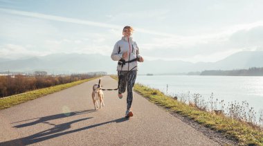Morning jogging with a pet: a female running together with her beagle dog by the asphalt way along lake with a foggy mountain landscape. Canicross exercises and active people and a dog concept image. clipart